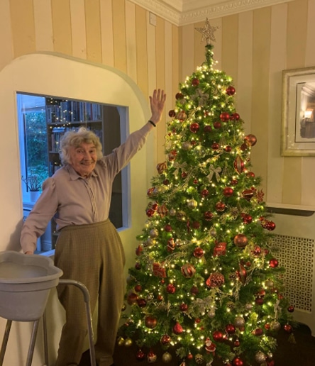 An elderly woman, in a beautifully decorated living room that's featured in numerous property listings, stands next to a christmas tree, reaching out towards the star on top.
